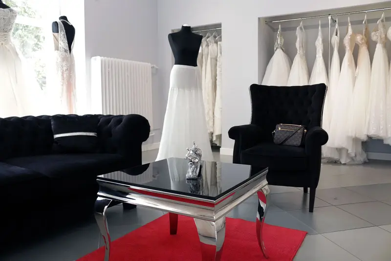 Salon Mija Loraa w Białymstoku: czarna pikowana sofa i fotel, lustrzany stolik na czerwonym dywanie, w tle suknie ślubne na wieszakach i manekinach.